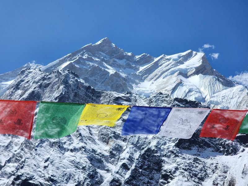 Colorful Tibetan prayer flags flutter in the wind against a backdrop of snow-covered Himalayan peaks, with Annapurna’s majestic, icy ridgelines rising under a clear blue sky.