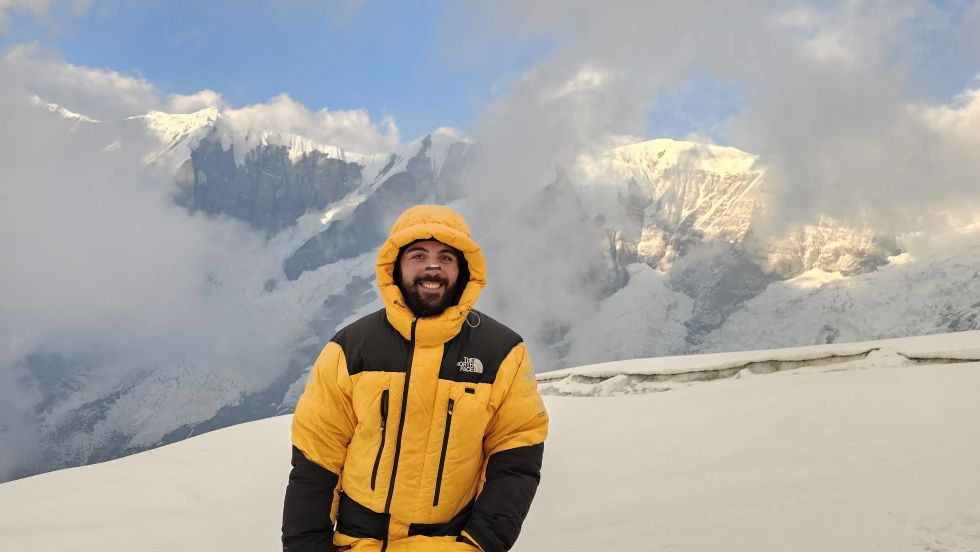 Zura Kvestadze in a yellow and black down jacket, standing on snowy terrain with Annapurna’s dramatic, cloud-covered peaks in the background.