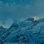 Snow-covered mountain peaks bathed in soft golden sunlight under a moody, cloud-filled sky.