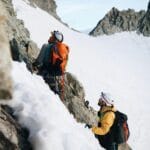Two mountaineers climb a steep, snowy, and rocky mountain face, roped together with a teal climbing rope. The lead climber, in a white helmet and gray jacket, carries an orange backpack, while the second climber wears a yellow jacket and white helmet with a black backpack. The background features snow-covered slopes and rugged peaks under a light blue sky with scattered clouds.