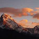 Annapurna I's snow-capped peaks glow orange at sunset, contrasting with dark slopes below. Peach-colored clouds fill the sky, framing the Himalayan giant's rugged beauty.