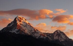 Annapurna I's snow-capped peaks glow orange at sunset, contrasting with dark slopes below. Peach-colored clouds fill the sky, framing the Himalayan giant's rugged beauty.
