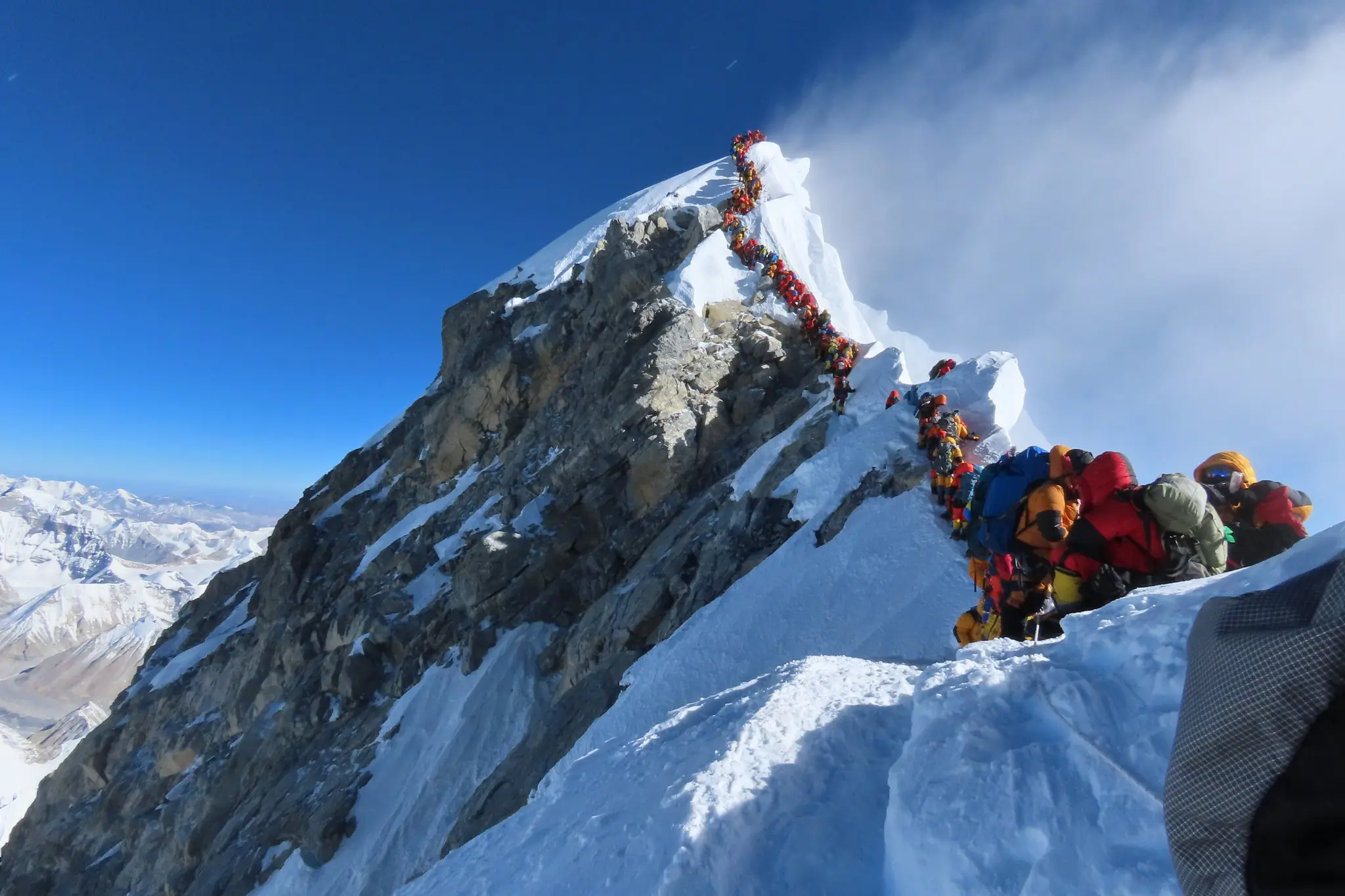 A long line of climbers in brightly colored gear ascends a narrow, snow-covered ridge near the summit of Mount Everest, highlighting overcrowding on the mountain against a backdrop of clear blue sky and distant Himalayan peaks.