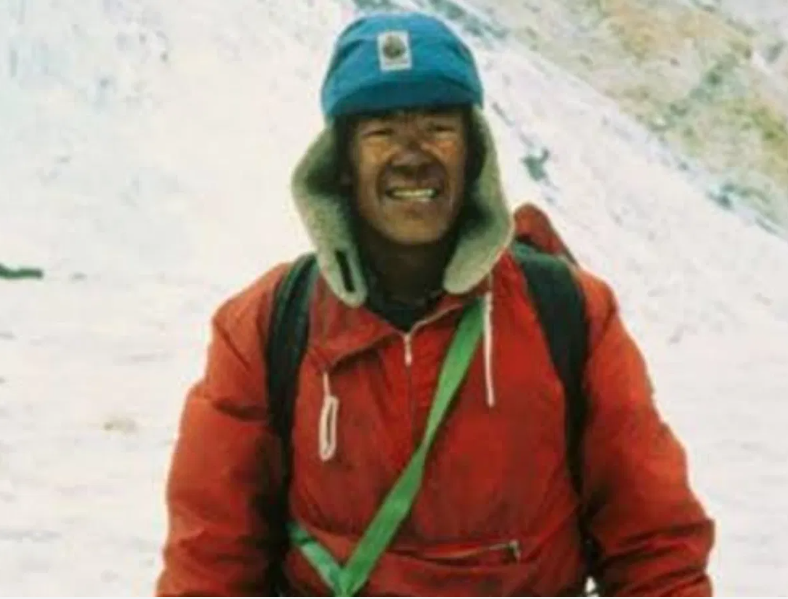 A mountaineer wearing a red jacket and blue hat with ear flaps stands smiling in a snowy mountain environment. He has a green strap across his chest and a backpack on his shoulders.
