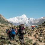 A group of hikers with backpacks walks along a rocky trail in a mountainous region, with a snow-capped peak dominating the background under a clear blue sky. Some porters carry large loads on their backs, and the landscape features sparse vegetation and rugged terrain.