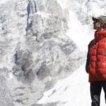 A man wearing a red and blue down jacket, black leggings, sunglasses, and a cap stands in front of a towering, snow-covered mountain face, looking upward. The rugged alpine backdrop suggests a high-altitude location, likely in the Himalayas.