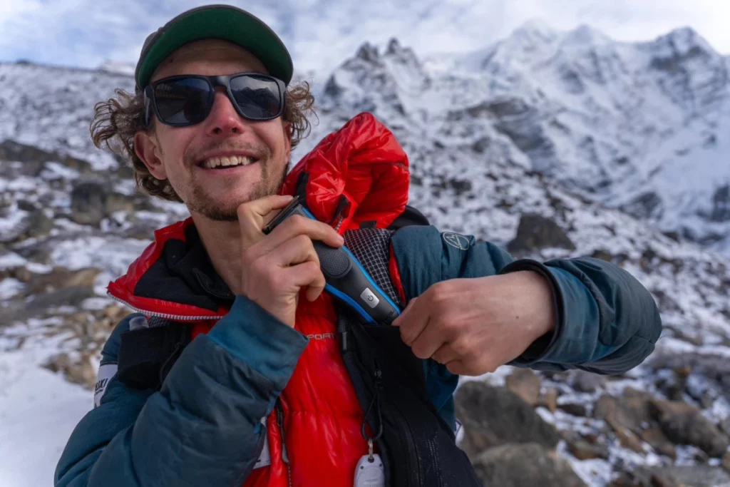 American endurance athlete Tyler Andrews smiles while holding a radio device to his chest in a snowy, mountainous environment. Dressed in a red and blue down jacket and wearing sunglasses and a green cap, he appears cheerful and energized, with rugged, snow-covered peaks rising in the background.








