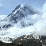 Snow-covered Mount Kazbek rises dramatically through a blanket of clouds, with the silhouette of the Gergeti Trinity Church visible on a hill in the foreground.
