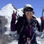 A smiling female mountaineer poses on a snowy mountain ridge, wearing a helmet, sunglasses, gloves, and climbing gear. She holds an ice axe in one hand and flashes a peace sign with the other, with snow-covered peaks and clear blue skies in the background