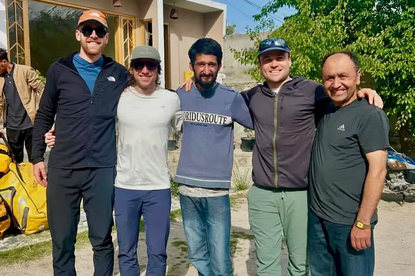 Five men standing side by side smiling in front of a house, including the Ultar Sar Southeast Pillar summit team and local supporters, enjoying a sunny day post-expedition.