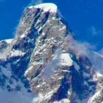 **Alt text:** Snow-covered Southeast Pillar of Ultar Sar towering sharply against a clear blue sky, with rugged rock faces and light clouds swirling around the flanks.
