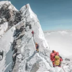 A group of climbers ascends a steep, snow-covered mountain ridge. They are roped together, wearing bright jackets and backpacks, with a clear blue sky above and clouds below, indicating a high altitude. The rocky, icy terrain is visible on both sides of the narrow path leading to the summit.