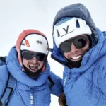 Two smiling climbers dressed in blue down suits and helmets take a selfie in snowy, high-altitude conditions. Both wear sunglasses and mountaineering gear, showing excitement and camaraderie during their expedition.