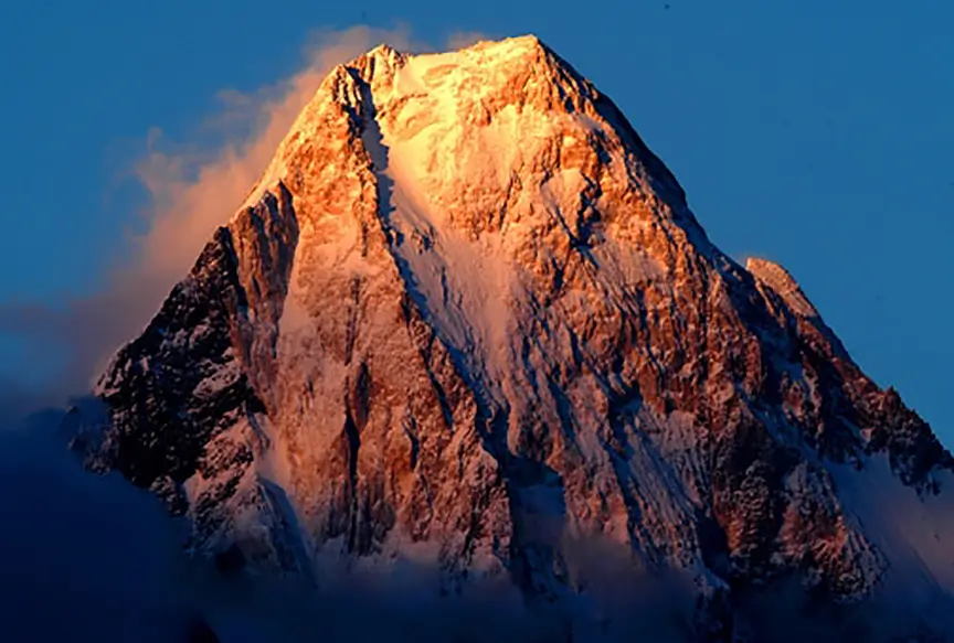 Gasherbrum IV's West Face—known as the Shining Wall—glows with golden alpenglow at sunset, revealing steep, rugged snow and rock lines against a clear, darkening sky. A dramatic and iconic high-altitude peak in the Karakoram.