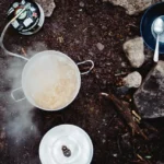 Overhead view of a camp cooking setup with a pot of boiling food on a portable gas stove, surrounded by rocks, a bowl with a spoon, food packaging, and outdoor gear on forest ground.