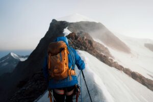 A skier with an orange helmet and backpack ascends a snow-covered slope, using ski poles, with rugged, snow-dusted mountains stretching into the distance under a cloudy sky.