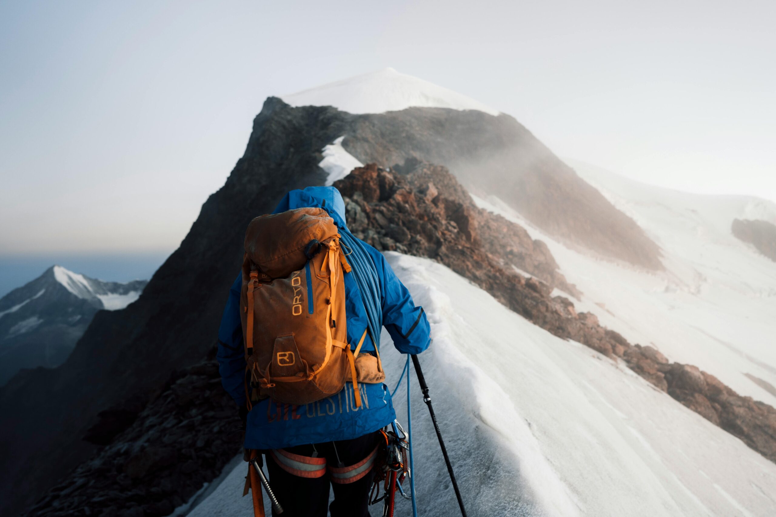 A skier with an orange helmet and backpack ascends a snow-covered slope, using ski poles, with rugged, snow-dusted mountains stretching into the distance under a cloudy sky.