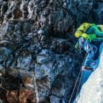A climber wearing bright gear and a yellow helmet ascends a steep icy rock face using ice axes and crampons, secured by ropes against a rugged cliff.