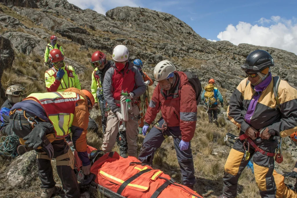 A group of mountain rescue team members, wearing helmets and climbing gear, tend to an injured person in an orange stretcher on a rocky hillside with a clear blue sky and distant peaks in the background.