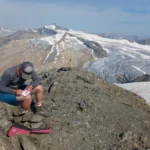 man is writing in notebook, background mountains