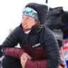 Laura Dahlmeier, dressed in a black and maroon insulated jacket with a TERREX logo, crouches in the snow smiling, wearing reflective ski goggles and a hood, during a winter sports event. A cameraman is visible in the blurred background.