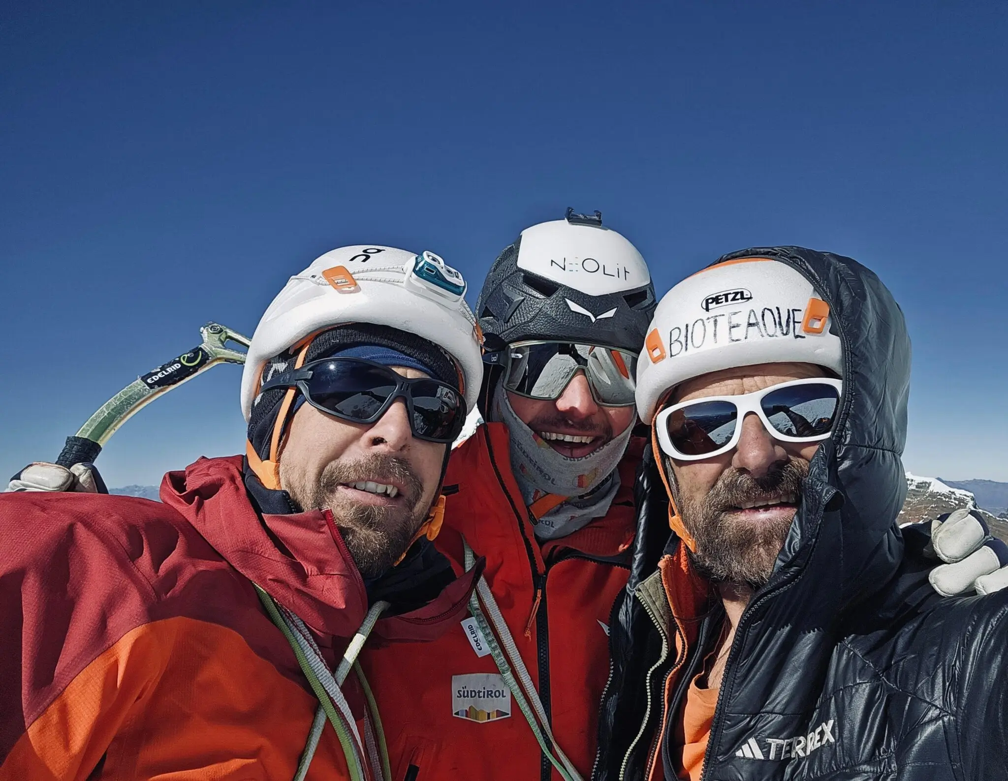 selfie of three smiling men on background of mountains, with helmets