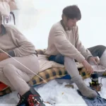 two men sit in base camp next to each other and are preparing food on gas, onr of the is waiting, one onf them prepares. wearing brown jackets