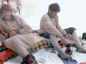 two men sit in base camp next to each other and are preparing food on gas, onr of the is waiting, one onf them prepares. wearing brown jackets