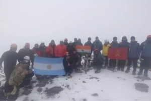 A group of people in winter gear posing together on a snowy mountain summit. They are holding several flags, including those of Argentina, India, and another with a red background and yellow symbol. The background is misty, with snow and rocks visible on the ground.