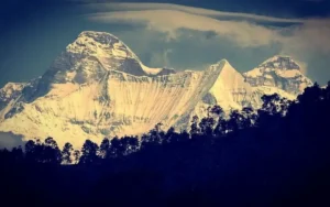 Snow‑covered twin peaks of Nanda Devi and Nanda Devi East rising above a forested ridge in Uttarakhand, India, under a dramatic sky