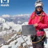 Alt text: A person wearing a bright pink jacket, climbing gear, and a white helmet with sunglasses stands on a snowy mountain peak. They hold a metal plaque with text, with a stunning backdrop of snow-covered mountains under a clear blue sky. The "mash" logo is visible in the upper left corner.