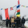 A group of climbers stands on a rocky, snow-covered mountain ridge, holding and surrounded by colorful prayer flags and national flags, including the Nepalese flag. The individuals are dressed in winter mountaineering gear, with some wearing sunglasses and hats. The background features a misty, mountainous landscape, suggesting a high-altitude location.