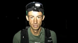 A person wearing a headlamp and a green shirt with the word "Normal" and a logo on it, standing against a dark background.