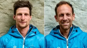 Two individuals wearing blue outdoor jackets with hoods, standing side by side against a stone wall background. The person on the left has short, dark hair and a slight smile, while the person on the right has short, light brown hair and a broader smile.