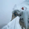 Two climbers are ascending a steep, icy mountain ridge. One climber in a red suit is positioned near the top of an ice wall, secured with ropes and climbing gear. Another climber in a blue suit is lower down, near a rocky outcrop, also roped up. The scene is covered in snow and ice, with icicles hanging from the rock face, and the background fades into a misty, cloudy sky.