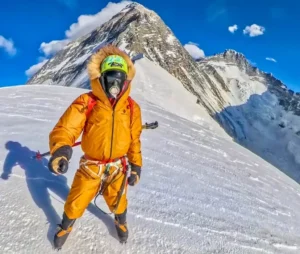A person wearing an orange mountaineering suit, gloves, and crampons stands on a snowy slope with ice axes in hand. They are equipped with a climbing harness and a green face mask with a respirator. In the background, a steep, snow-covered mountain peak rises under a clear blue sky with a few clouds.