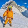 A person wearing an orange mountaineering suit, gloves, and crampons stands on a snowy slope with ice axes in hand. They are equipped with a climbing harness and a green face mask with a respirator. In the background, a steep, snow-covered mountain peak rises under a clear blue sky with a few clouds.