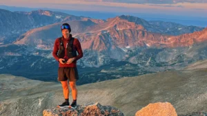 A person standing on a rocky peak with a scenic view of rugged mountains at sunset, wearing a red jacket, black shorts, and a headband, with a backpack and a device in hand.