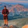 A person standing on a rocky peak with a scenic view of rugged mountains at sunset, wearing a red jacket, black shorts, and a headband, with a backpack and a device in hand.