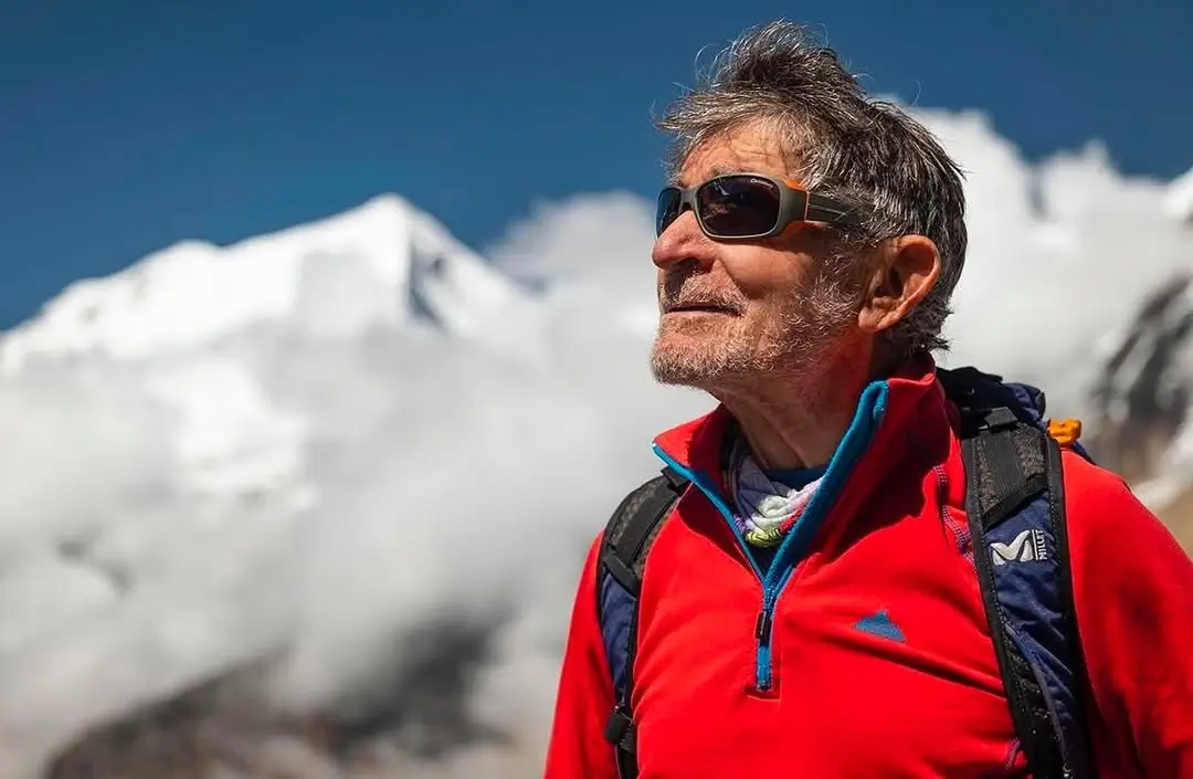 A person wearing a red jacket and sunglasses stands outdoors with a backpack, gazing upward. Snow-capped mountains and a clear blue sky with clouds form a stunning backdrop.