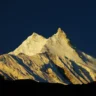 A stunning view of a snow-covered mountain peak illuminated by golden sunlight against a deep blue sky, with rugged slopes and shadows creating a dramatic contrast.