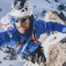 A climber wearing a white helmet with "SIMOND" branding and a blue jacket with "The North Face" logo ascends a rocky, snow-covered mountain, gripping the terrain with gloved hands and supported by climbing gear.