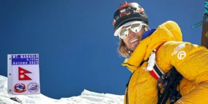 A person in a bright yellow mountaineering jacket and helmet stands on a snowy ridge with a clear blue sky in the background. To the left, a sign reads "MT. MANASLU 8,163m AUTUMN 2025" with the Nepalese flag and a logo of the Nepal Mountaineering Association. The person is equipped with climbing gear, including ropes and a harness.