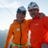 Two climbers wearing orange jackets and helmets stand with arms around each other, smiling, against a backdrop of mountains under a clear sky with a wooden pole and cables nearby.