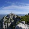 A rugged mountain peak with a cross at the summit, surrounded by rocky terrain and green shrubs. The background features a vast range of rolling hills and distant snow-capped mountains under a clear blue sky with wispy clouds.