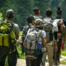A group of hikers walking along a narrow trail in a lush green forest, each carrying large backpacks, with dense vegetation and rocky terrain on either side.