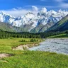 A scenic landscape featuring a lush green valley with a winding river, surrounded by dense pine forests and vibrant wildflowers. In the background, a range of snow-capped mountains rises sharply under a clear blue sky with scattered clouds. A dirt path cuts through the meadow, adding to the natural beauty of the scene.