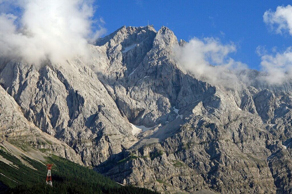 A dramatic snow-capped alpine peak towers above jagged rocky ridges under a deep blue sky. Patches of snow cling to the steep granite faces, with lower slopes revealing hints of green vegetation and distant mountain ranges fading into the horizon.