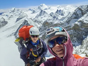 Two climbers taking a selfie on a snowy mountain ridge, with stunning views of snow-covered peaks under a clear blue sky. Both are wearing helmets, sunglasses, and climbing gear, with one carrying a large orange backpack and the other a camera attached to their helmet.