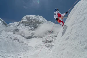 A climber in a red and white suit and blue helmet ascends a steep, snow-covered mountain slope, secured with orange climbing ropes. The rugged, icy peak and surrounding snow-covered ridges rise sharply against a clear blue sky, with a faint rainbow visible in the distance.